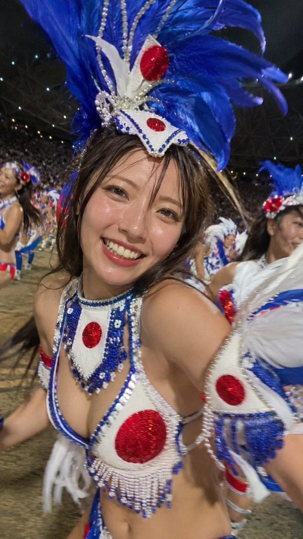 A smiling woman in a blue and white carnival-style costume with red accents, resembling the Japanese flag, poses for a selfie at night.