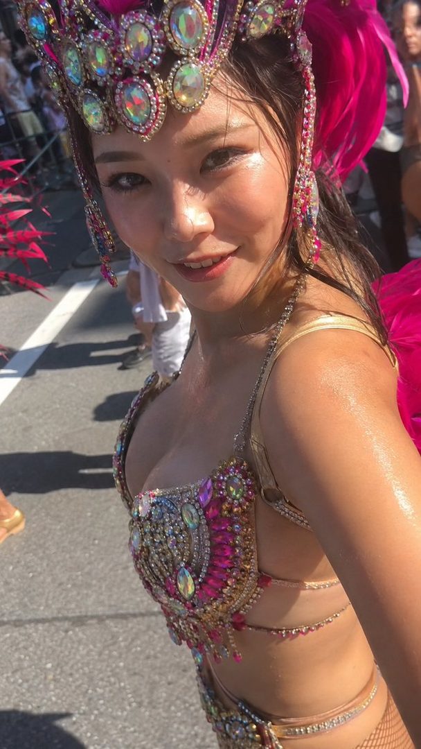 A woman in a vibrant, jeweled carnival costume with pink feathers smiles at the camera on a sunny street.
