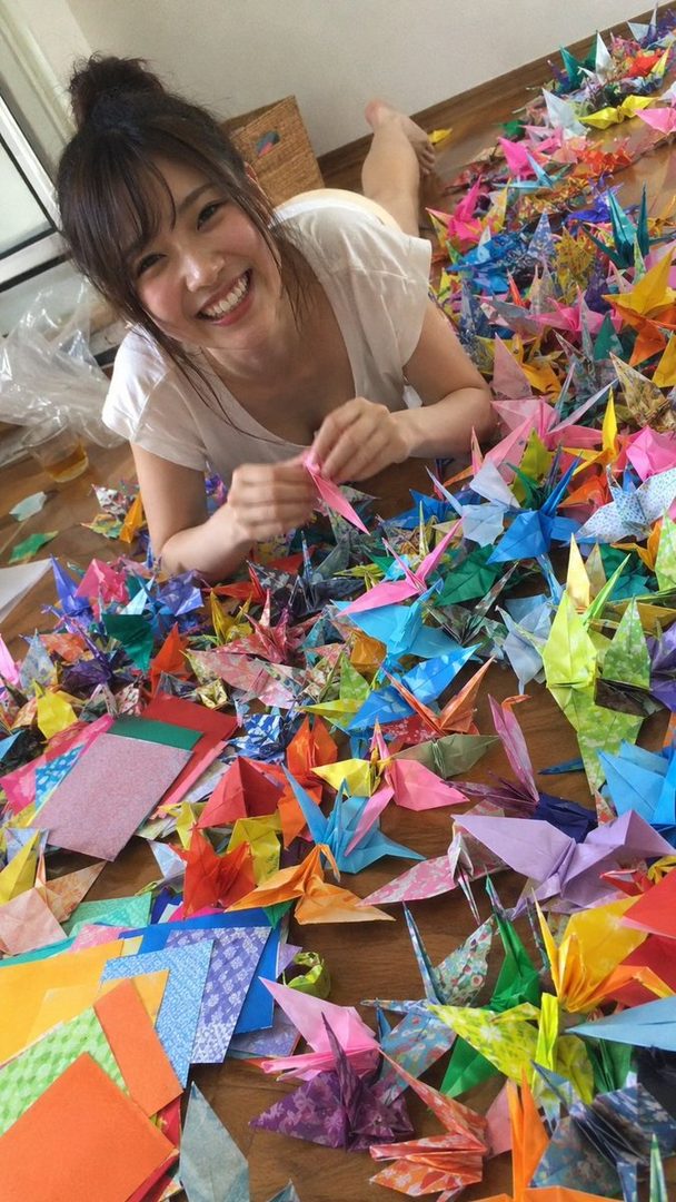 A smiling woman lies on the floor surrounded by colorful origami cranes, holding one in her hands.