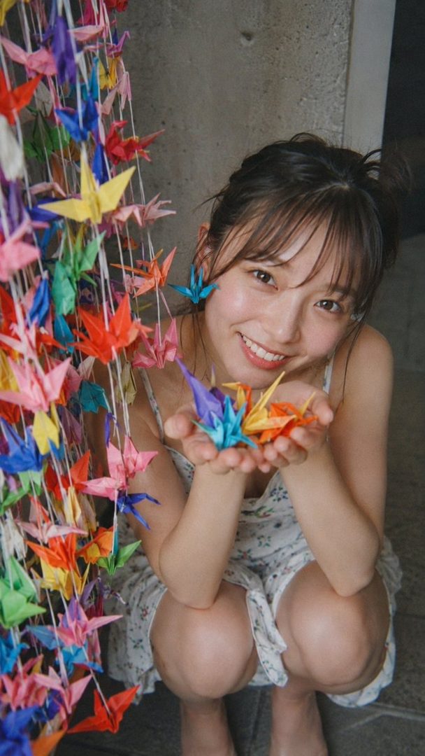 A young woman in a floral dress crouches and smiles, holding colorful origami cranes in her hands, with more origami cranes hanging beside her.
