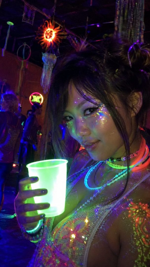 A woman with glitter makeup and glow-in-the-dark accessories holds a glowing cup in a dark, neon-lit club.