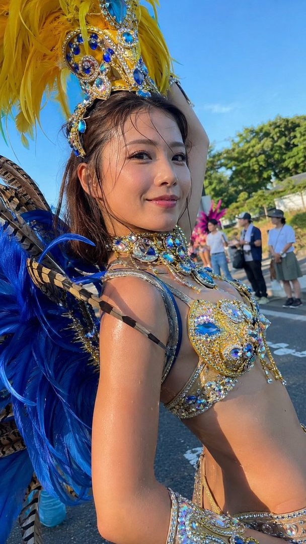 A woman in a vibrant samba costume with blue and gold feathers and jewels smiles at the camera on a sunny street.