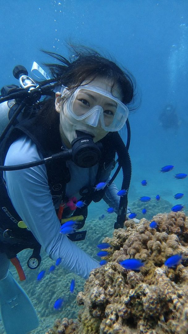 A Japanese woman in scuba gear smiles at the camera underwater, surrounded by small blue fish and coral.