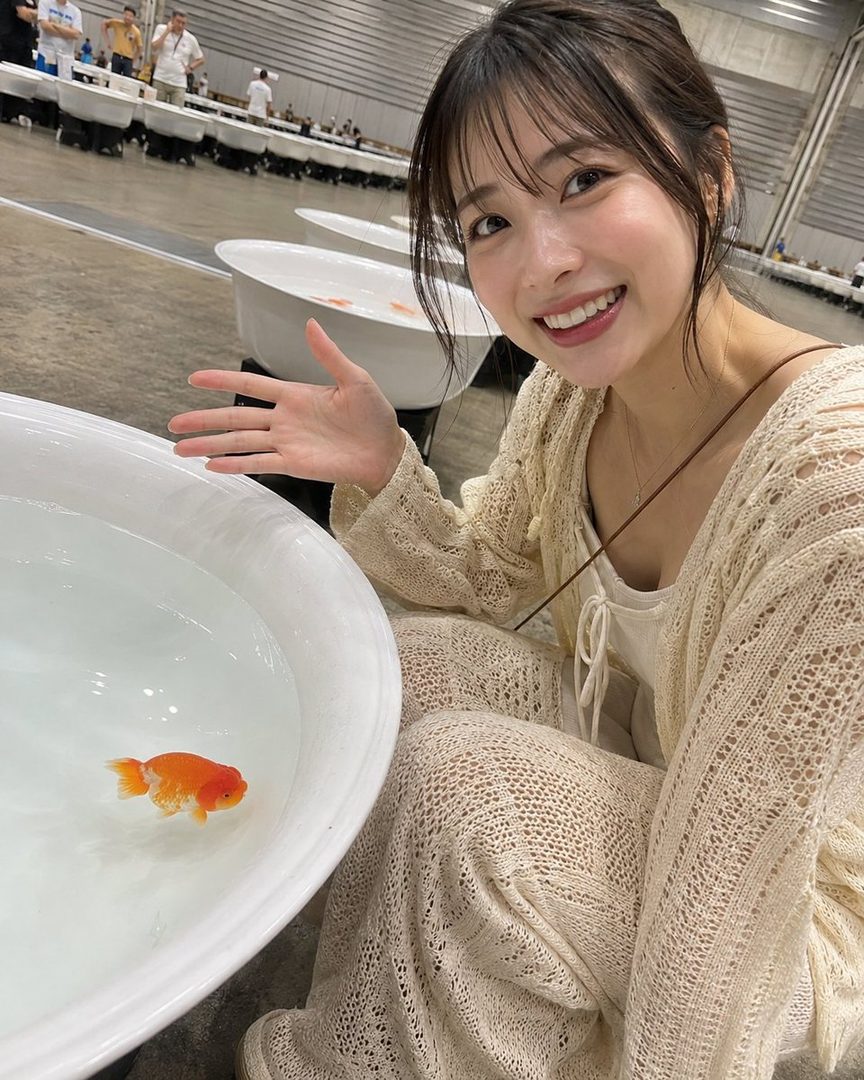 A smiling young woman with brown hair and bangs, wearing a light-colored knit top, poses next to a white bowl with a goldfish in it, indoors.