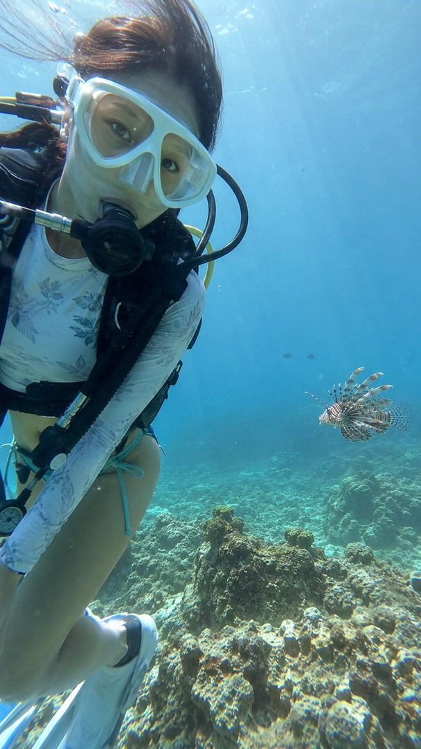 A woman in scuba gear swims underwater near a coral reef, with a lionfish in the background.