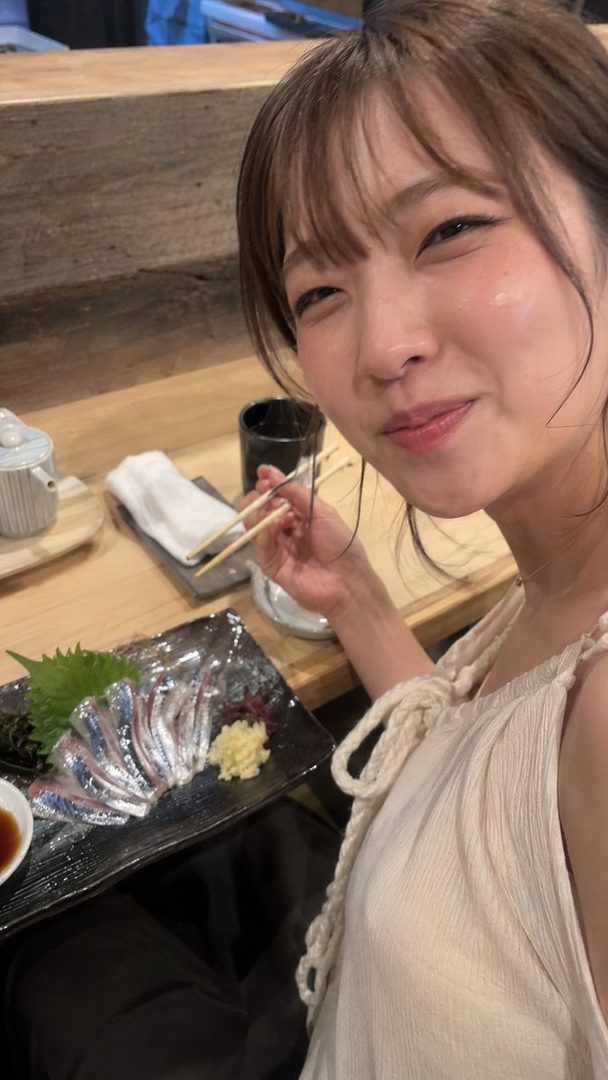 A young woman with brown hair and bangs smiles while holding chopsticks over a plate of sashimi at a restaurant.