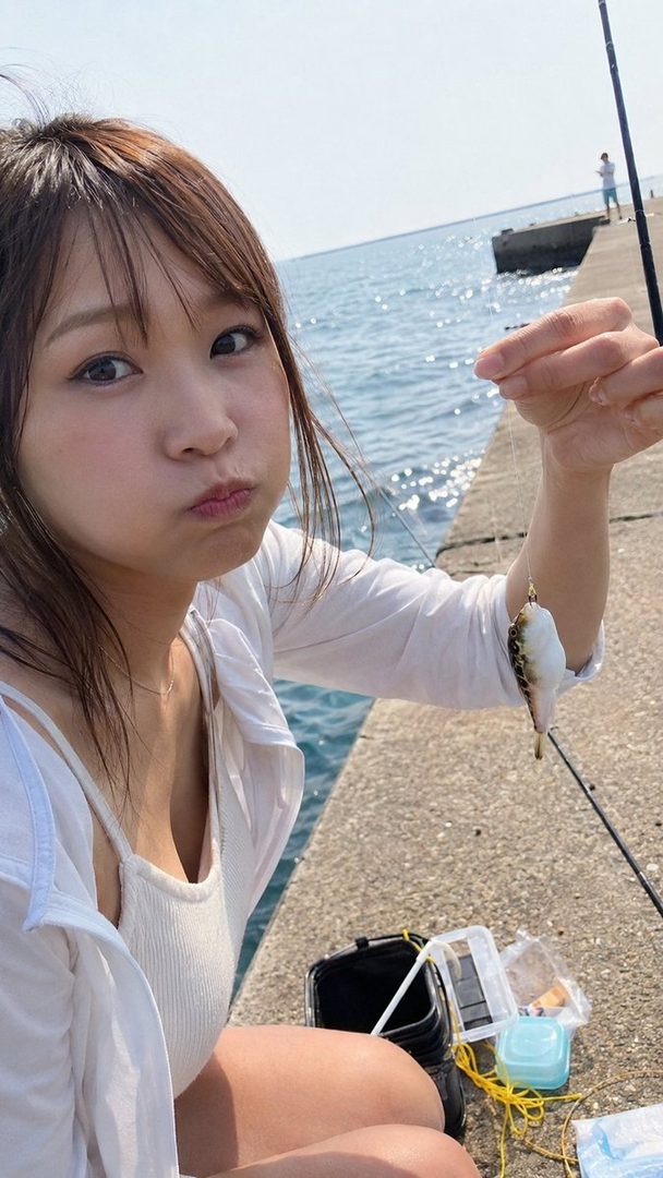 A young woman with brown hair pouts while holding a small fish on a fishing line by the sea.