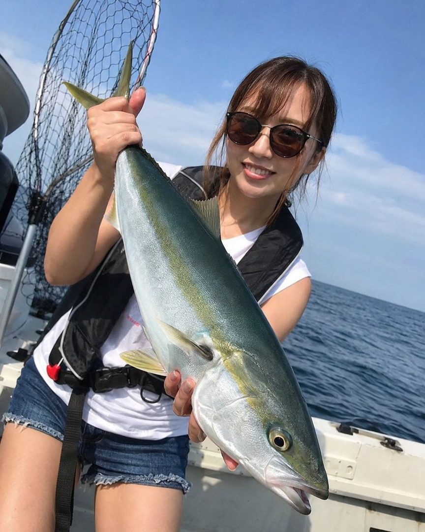 A woman wearing sunglasses and a life vest holds up a large fish on a boat under a clear blue sky.