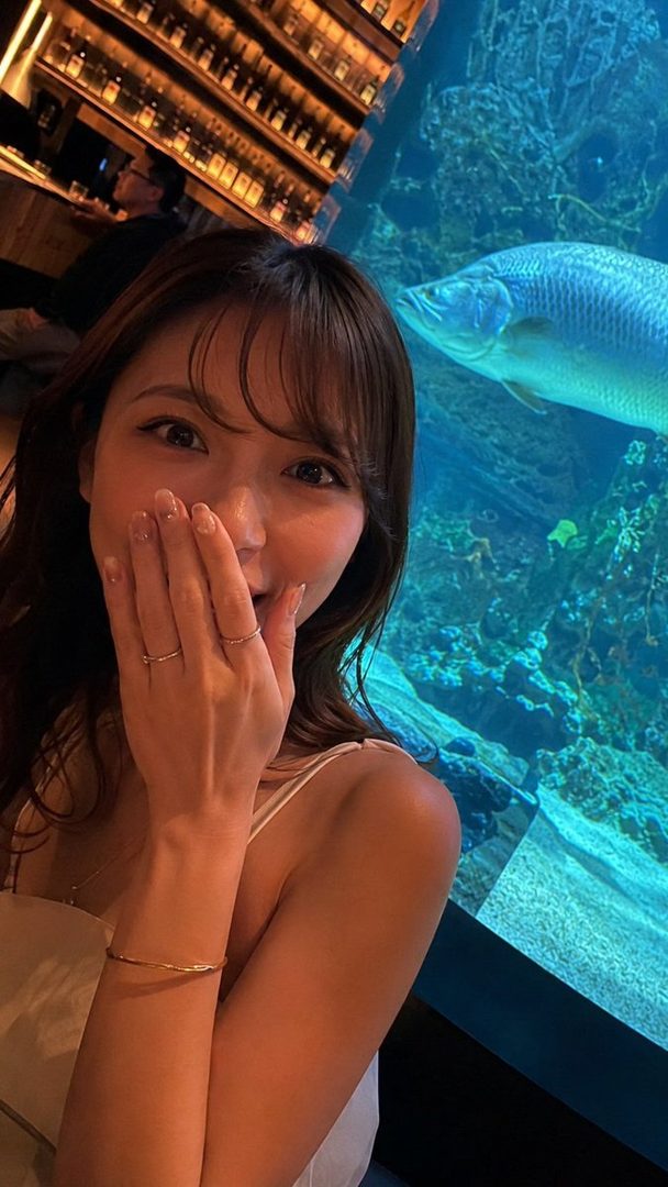 A young woman takes a selfie indoors, covering her mouth with her hand, with an aquarium and a bar in the background.