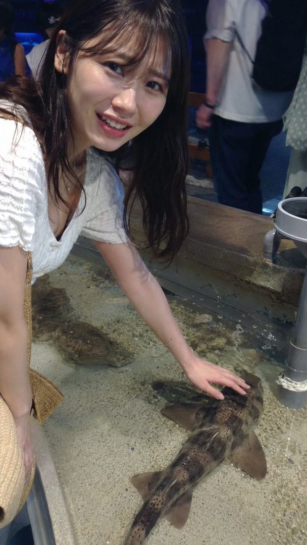 A young woman with long dark hair and bangs smiles while gently touching a shark in an indoor aquarium.
