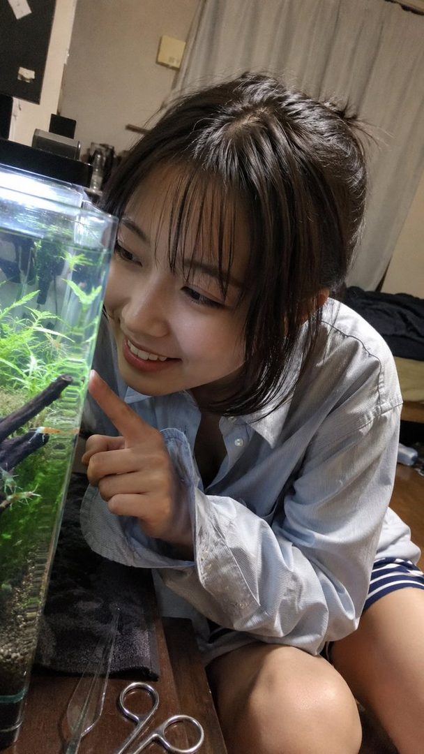 A young woman with bangs and a casual shirt smiles while pointing at an aquarium with aquatic plants.