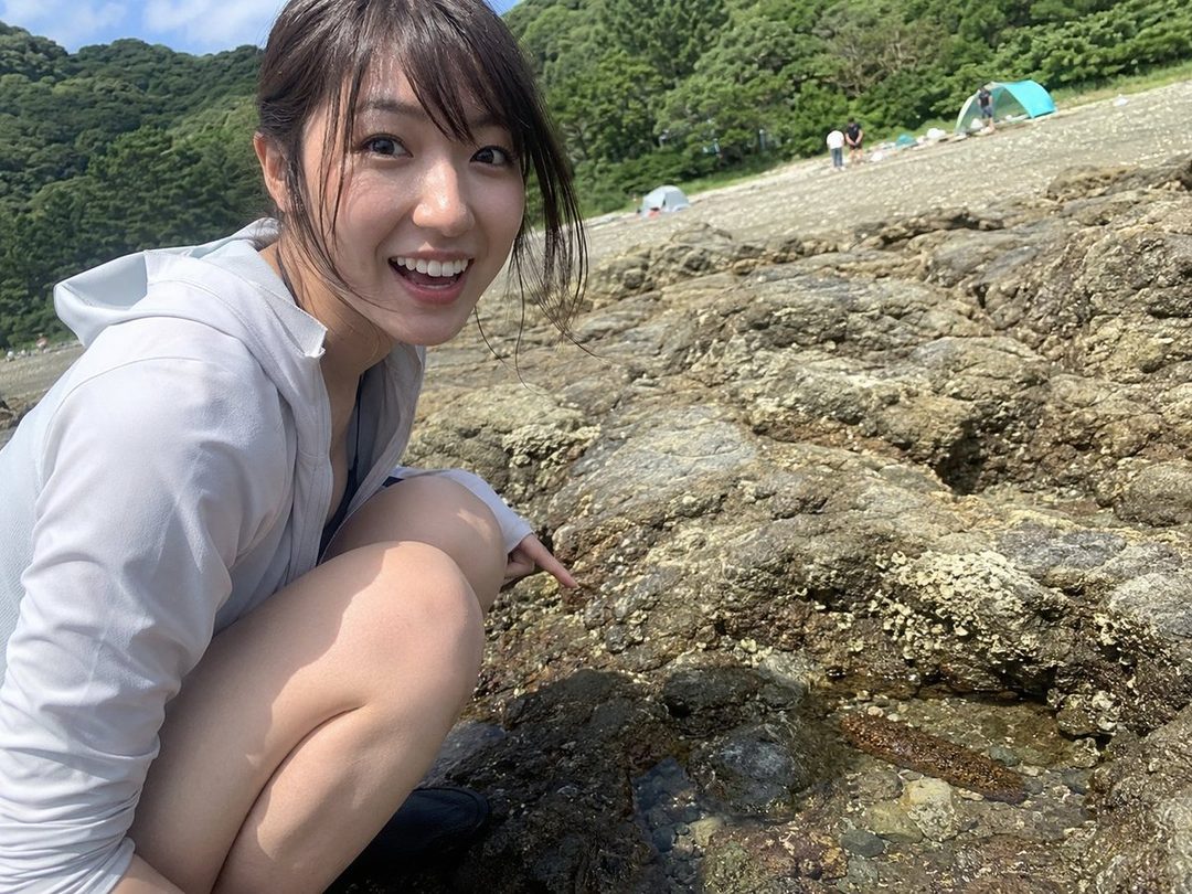 A young woman in a white hoodie crouches on a rocky beach, pointing at a sea cucumber in a tide pool, with a bright smile.