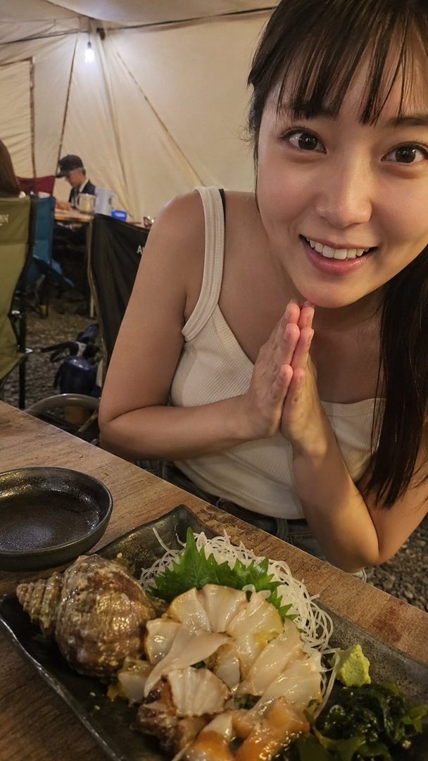 A young woman with dark hair and bangs smiles at the camera, holding her hands together in a gesture of gratitude, with a plate of seafood in front of her at an indoor dining setting.