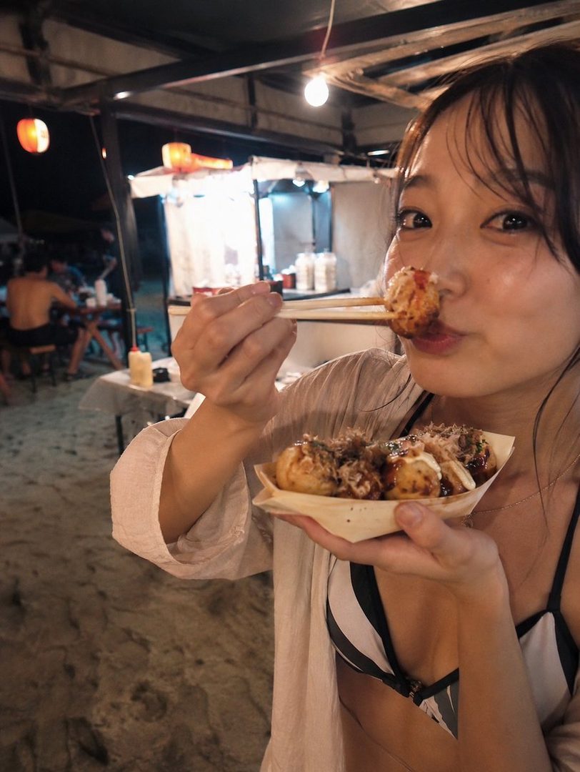 A young woman in a swimsuit and light cover-up playfully holds takoyaki with chopsticks, with a street food stall in the background at night.