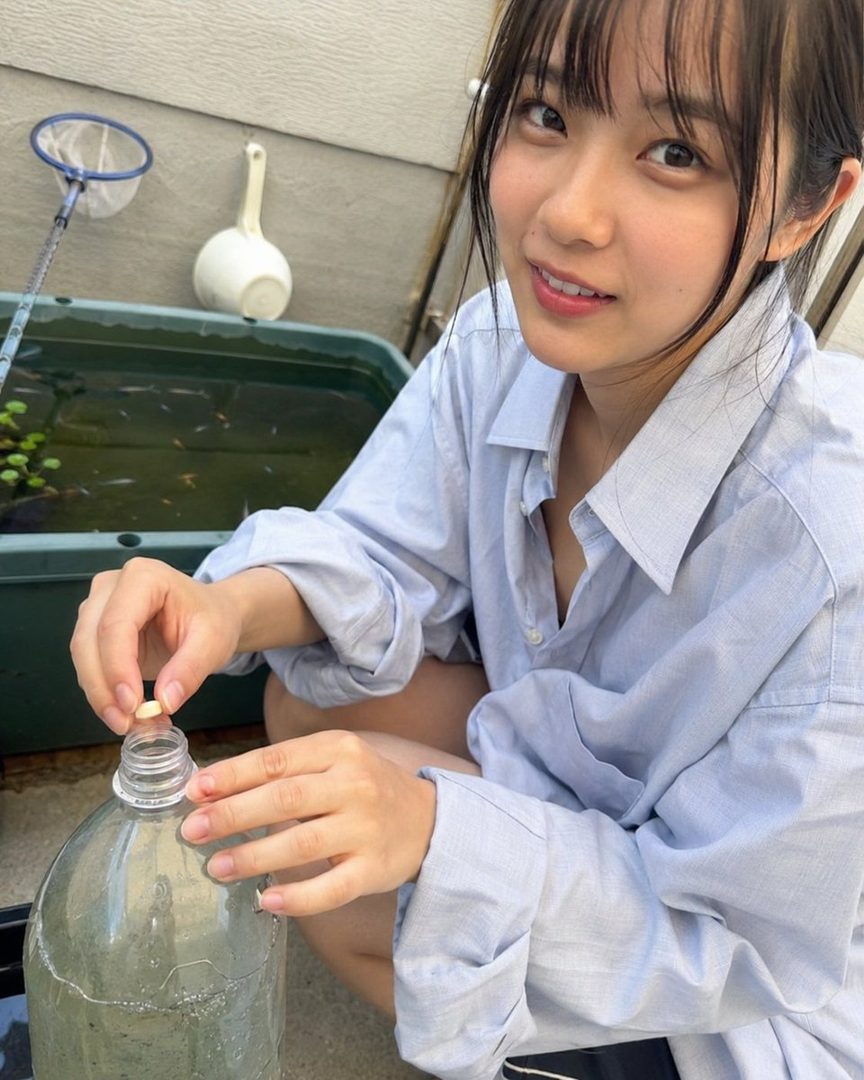 A young woman with black hair and bangs, wearing a light blue shirt, is crouching outdoors and putting something into a plastic bottle, with a small fish tank in the background.