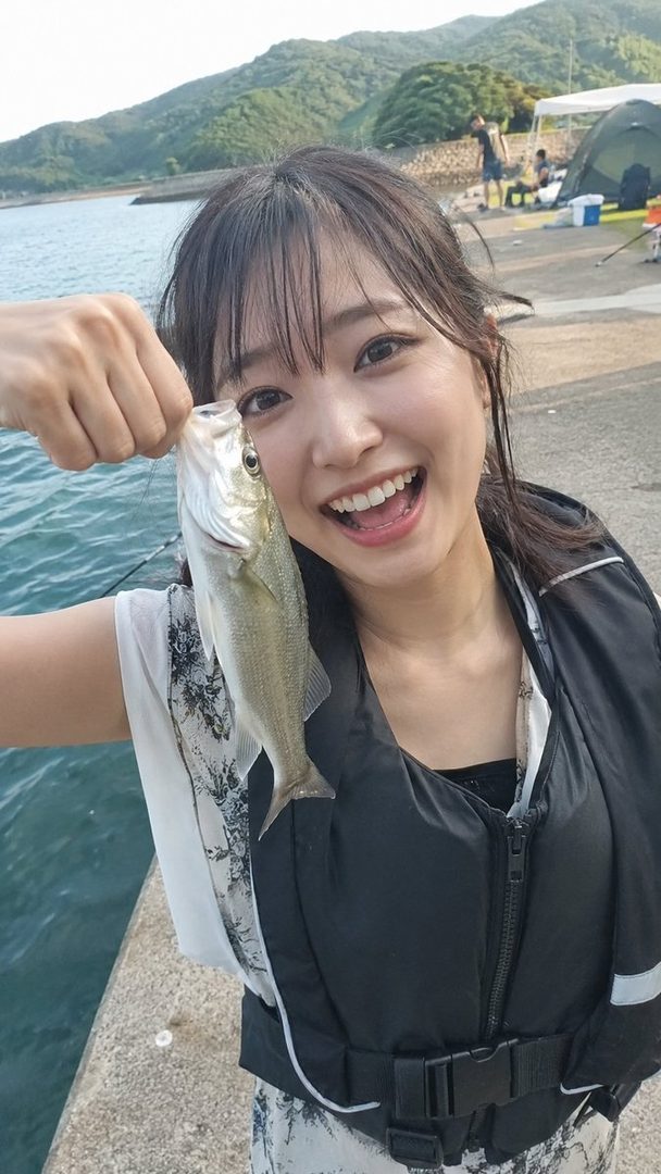 A smiling young woman in a life vest holds up a small fish she caught, with a scenic outdoor background of water and mountains.