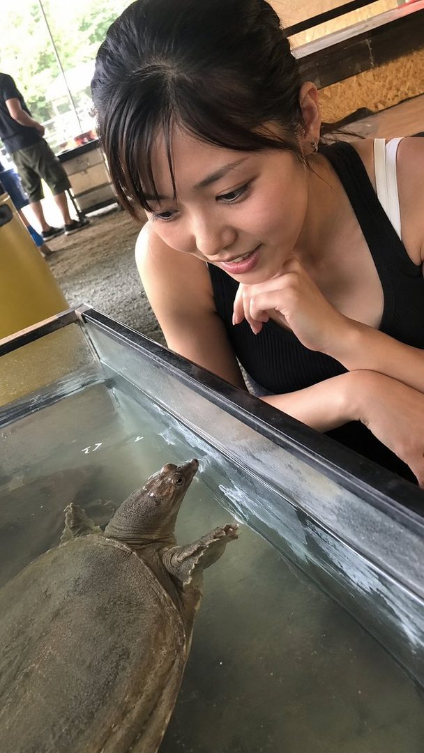 A young woman with black hair and bangs, wearing a black tank top, looks down at a turtle in a tank.