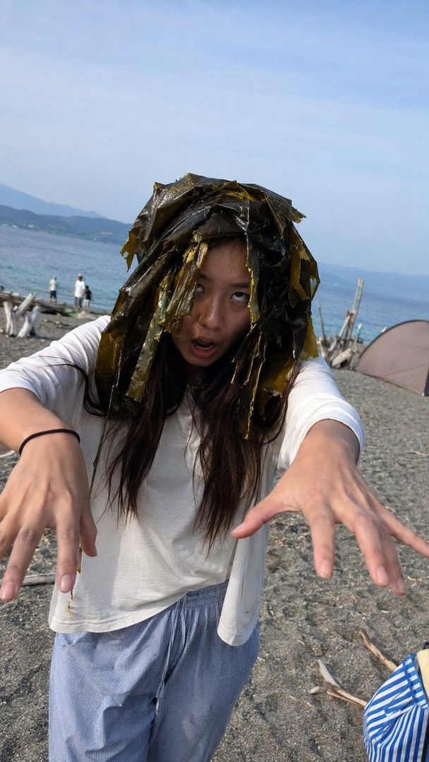 A woman with seaweed on her head makes a funny face and extends her hands towards the camera on a beach.