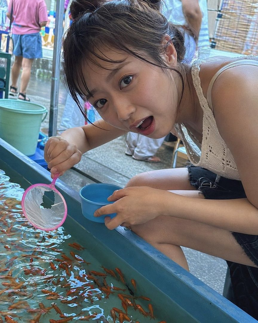 A young woman with brown hair tied up, wearing a white top and black shorts, is playing goldfish scooping at an outdoor festival.