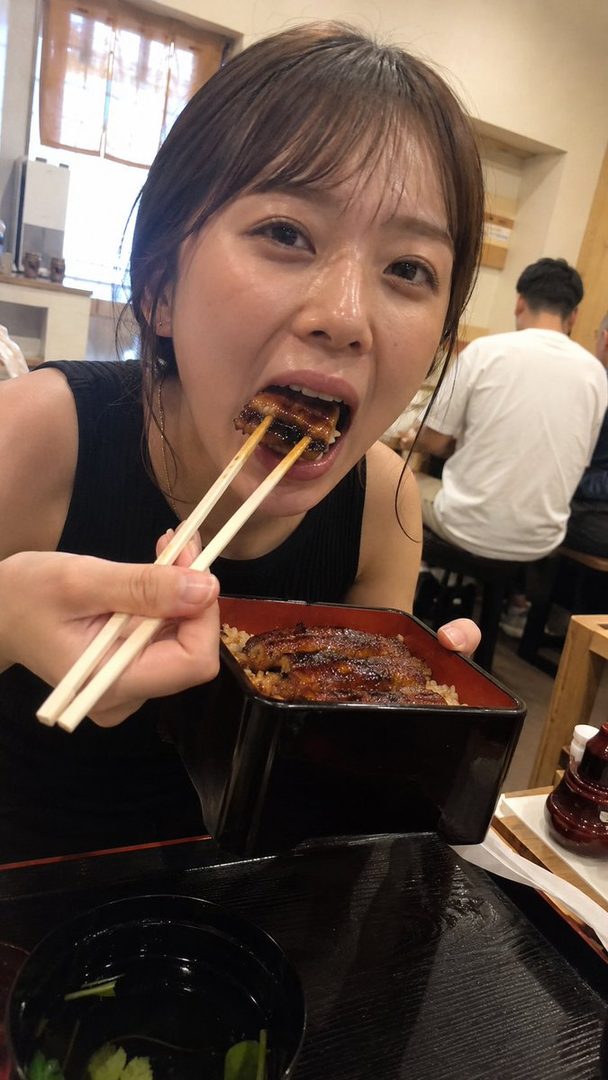 A young woman with brown hair and bangs is captured mid-bite, eating unagi (grilled eel) from a black bento box with chopsticks in an indoor setting.