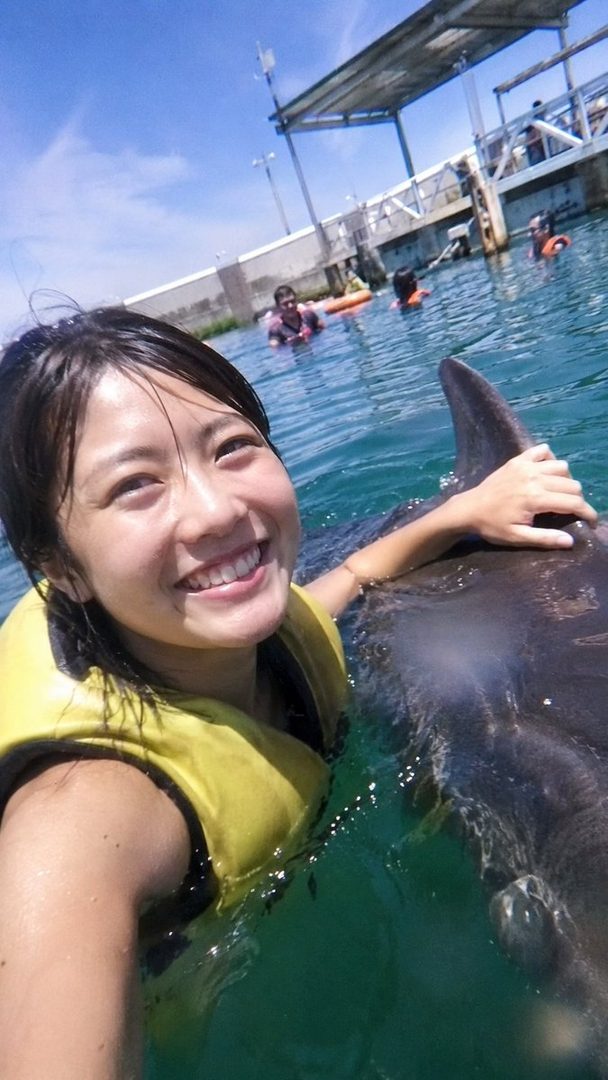 A smiling woman in a yellow life vest takes a selfie while swimming with a dolphin in clear blue water.