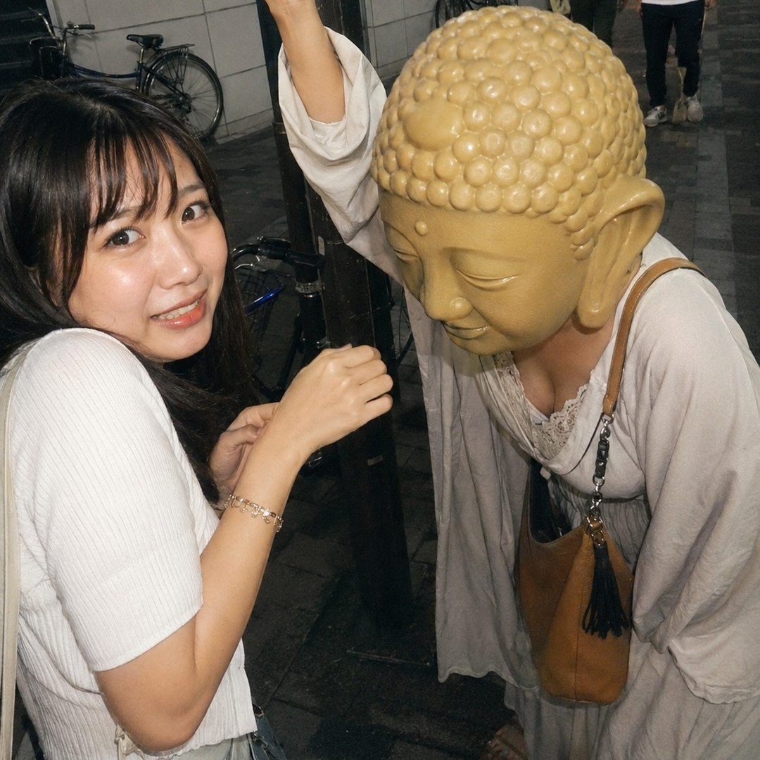 A young woman with long black hair and bangs smiles at the camera next to a person wearing a Buddha head mask on a street at night.