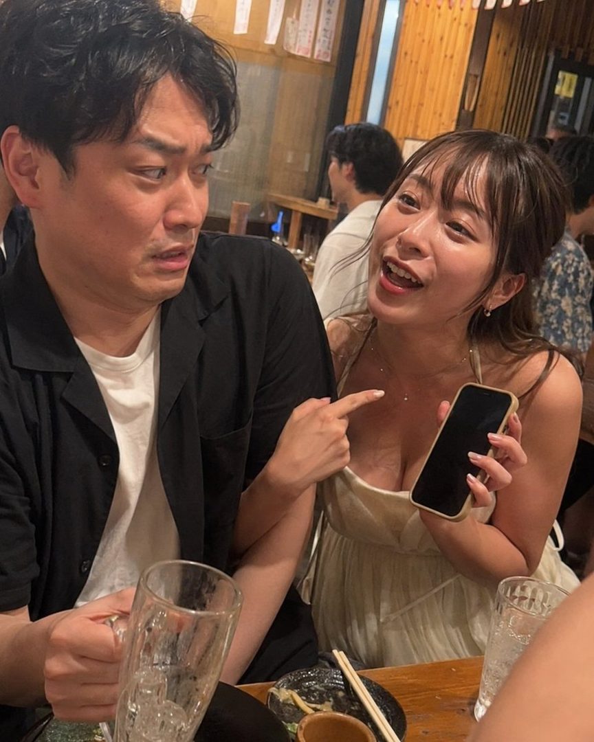 A woman in a light dress points at her chest while holding a phone, looking at a man with a surprised expression in an indoor setting.