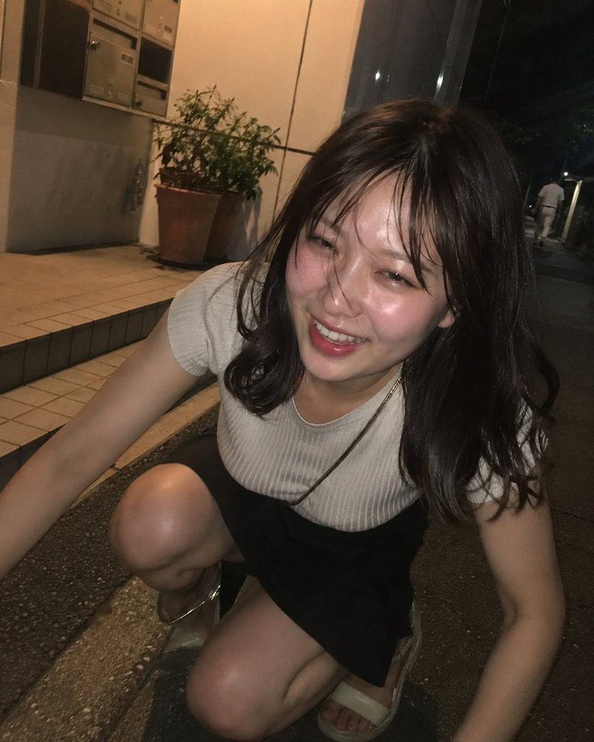 A young woman with long brown hair, wearing a light top and dark skirt, crouches on a street at night, smiling brightly.