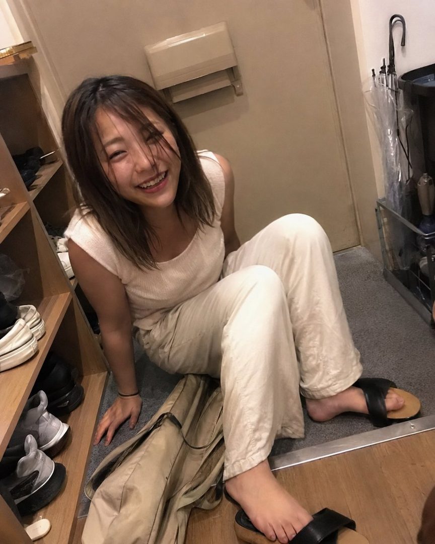 A young Japanese woman with short brown hair, wearing a light-colored top and pants, sits on the floor and smiles brightly at the camera.