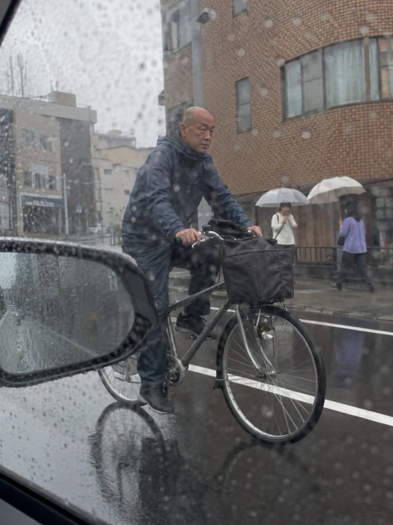 A man in a dark raincoat rides a bicycle on a wet street during a rainy day, with buildings and other pedestrians in the background.