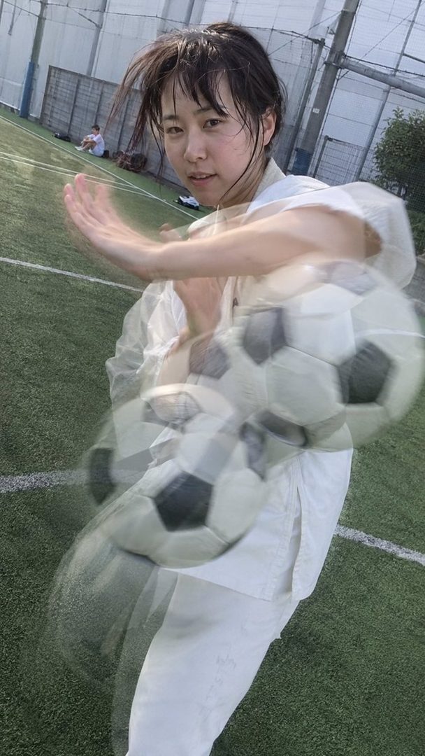A young woman in sportswear is captured in motion on a soccer field, with multiple soccer balls blurred around her, suggesting dynamic movement.