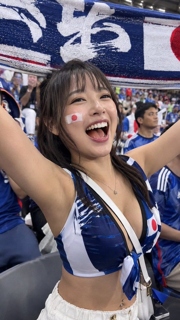 A young woman with a Japanese flag painted on her cheek cheers enthusiastically at a sports event, wearing a blue and white crop top.