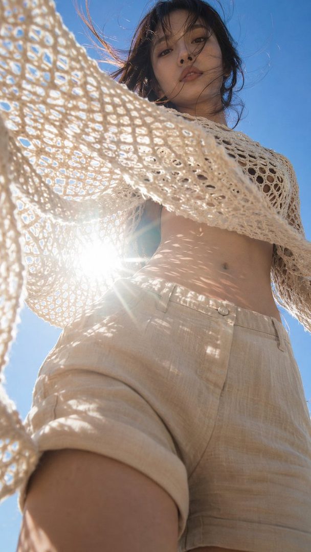 A young woman with brown hair and bangs wears a crocheted top and shorts, with sunlight flaring behind her against a clear blue sky.