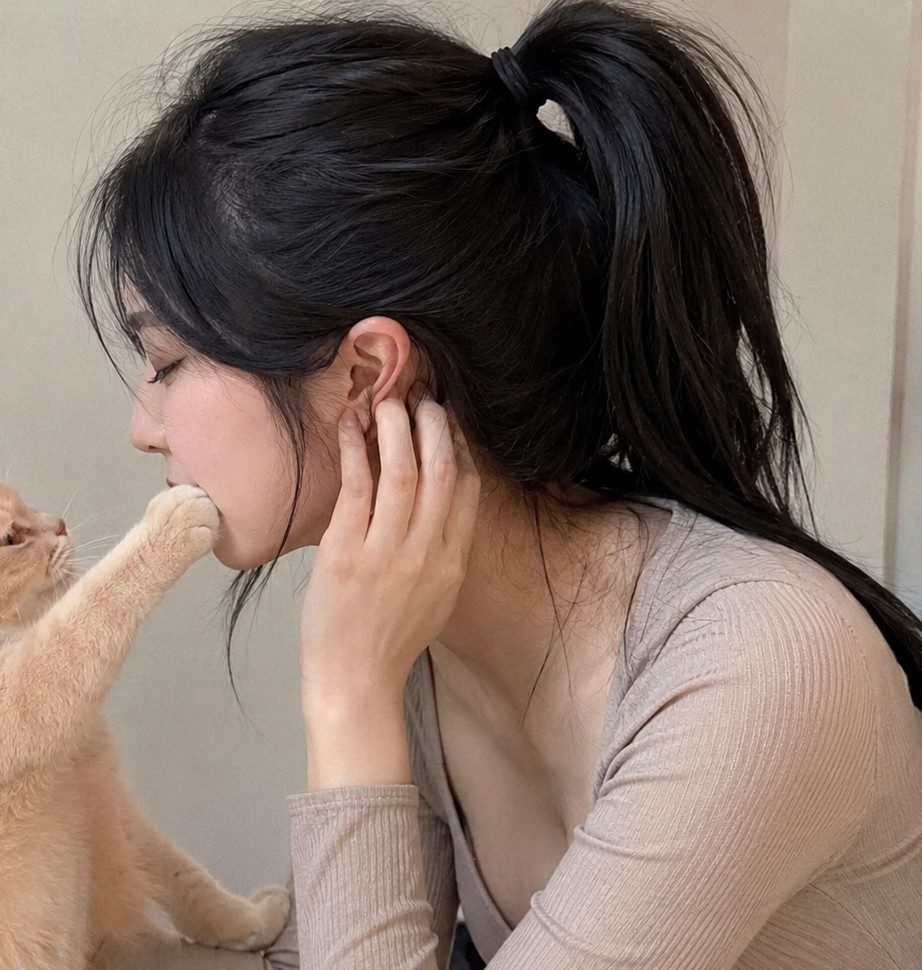 A woman with black hair in a ponytail is shown in profile, with a cat's paw gently touching her mouth.
