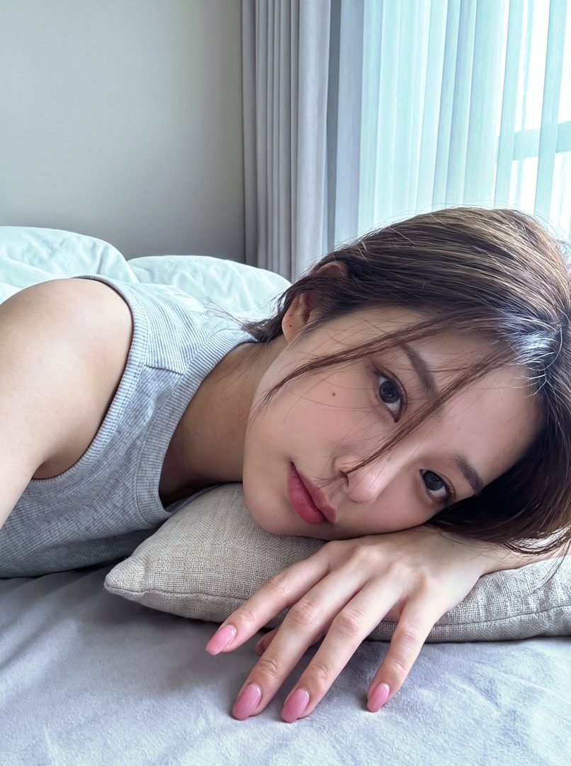 A photorealistic portrait of a young woman with short brown hair, wearing a grey tank top, lying on a bed with her head on a pillow, looking at the camera.