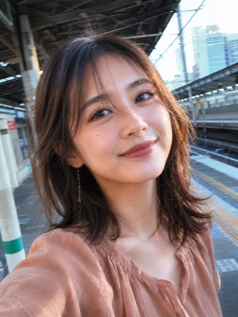 A young woman with brown hair takes a selfie on a train platform, smiling softly at the camera.