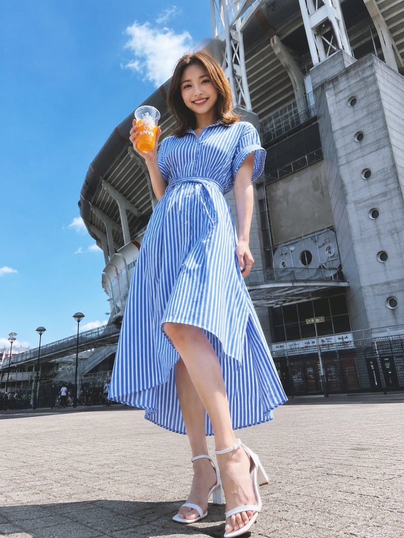 A smiling woman in a blue and white striped dress holds a drink while standing outdoors in front of a large building.