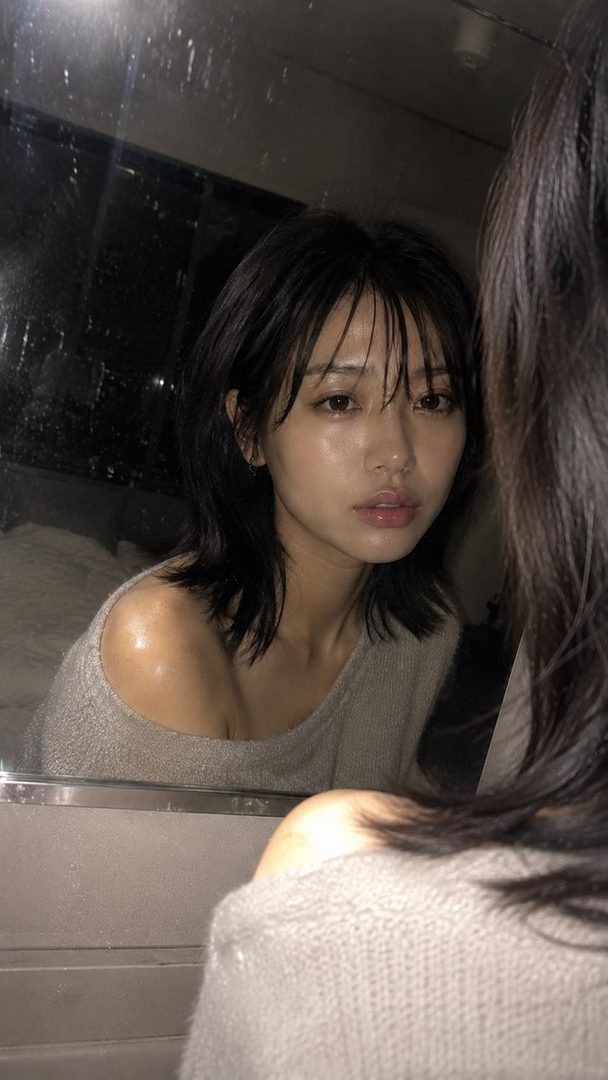 A woman with short black hair and bangs looks into a mirror, wearing an off-the-shoulder knit top.