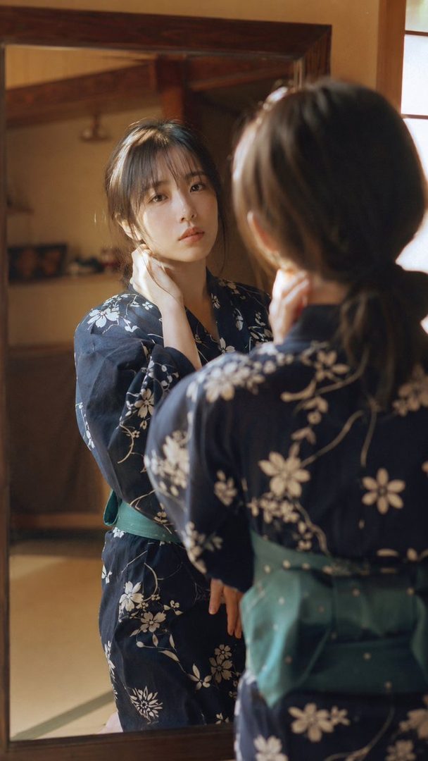 A young woman in a dark blue yukata with white floral patterns looks at her reflection in a mirror.
