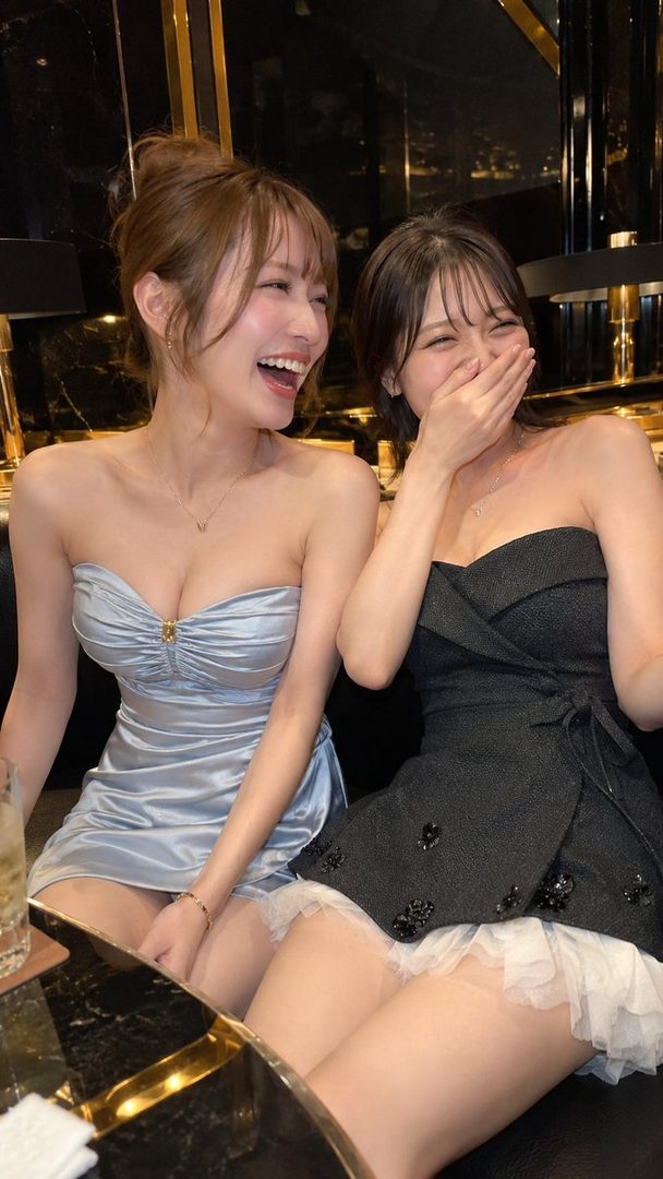 Two women in strapless dresses are laughing together at a table in a dimly lit indoor setting.