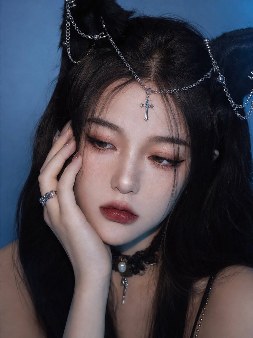 A close-up portrait of a woman with long black hair, wearing cat ears with silver chains and a cross pendant, and a black lace choker, against a blue background.