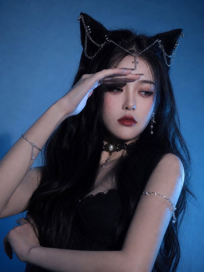 A woman with long black hair and cat ear headband poses against a blue background, shielding her eyes with her hand.