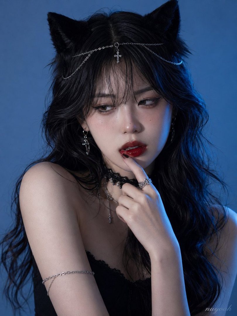 A photorealistic portrait of a woman with long black hair and cat ears, wearing dark makeup and a black top, against a blue background.