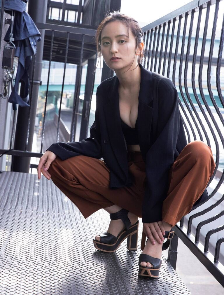 A Japanese woman in a black blazer and brown pants squats on a metal platform outdoors, looking directly at the camera.