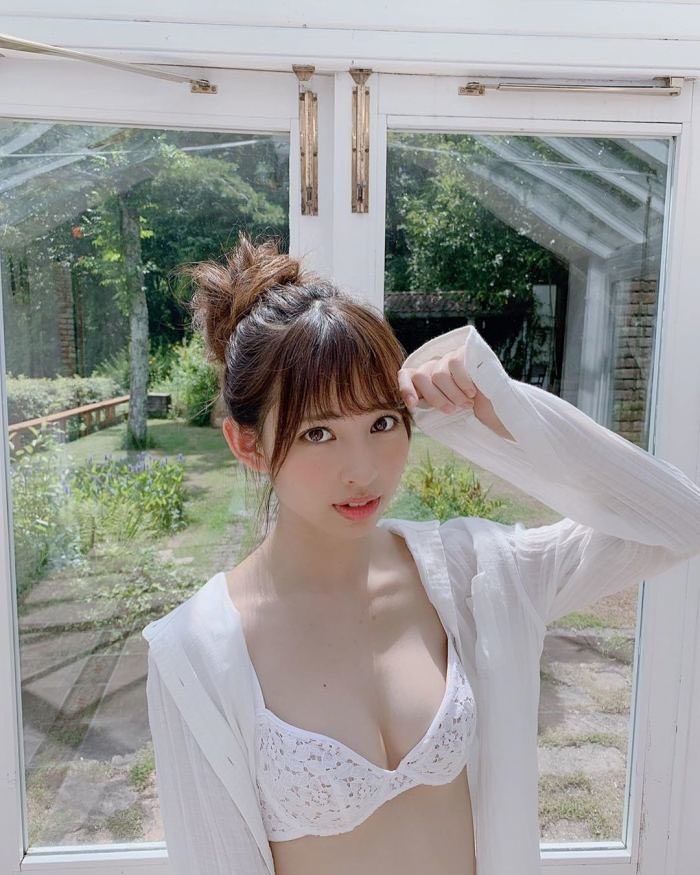 A Japanese woman with brown hair in a bun and bangs, wearing a white lace bra and a sheer white cardigan, poses indoors with a garden visible through glass doors.