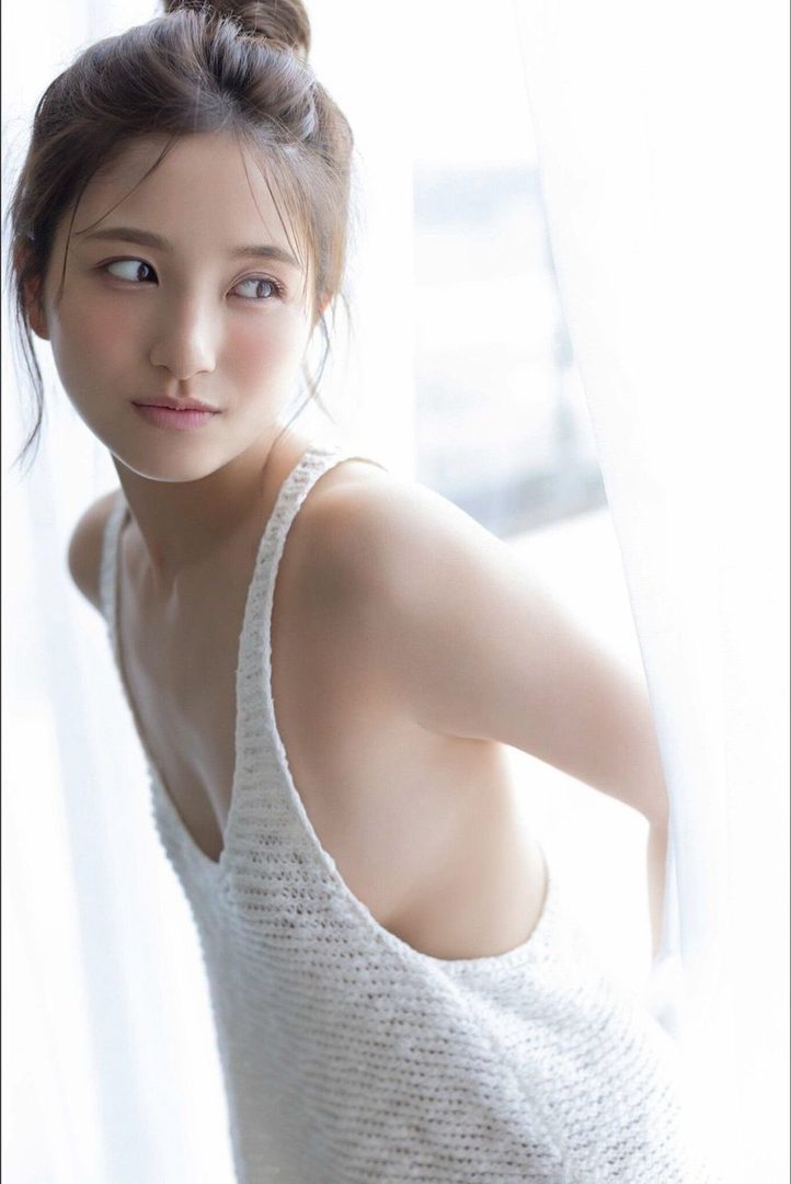 A young Japanese woman with her hair in a bun, wearing a white knit top, looks away from the camera in a brightly lit indoor setting.