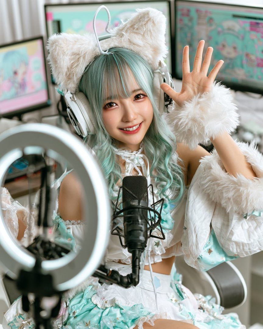 A smiling woman with light blue hair and cat-ear headphones poses in front of a microphone and computer monitors.