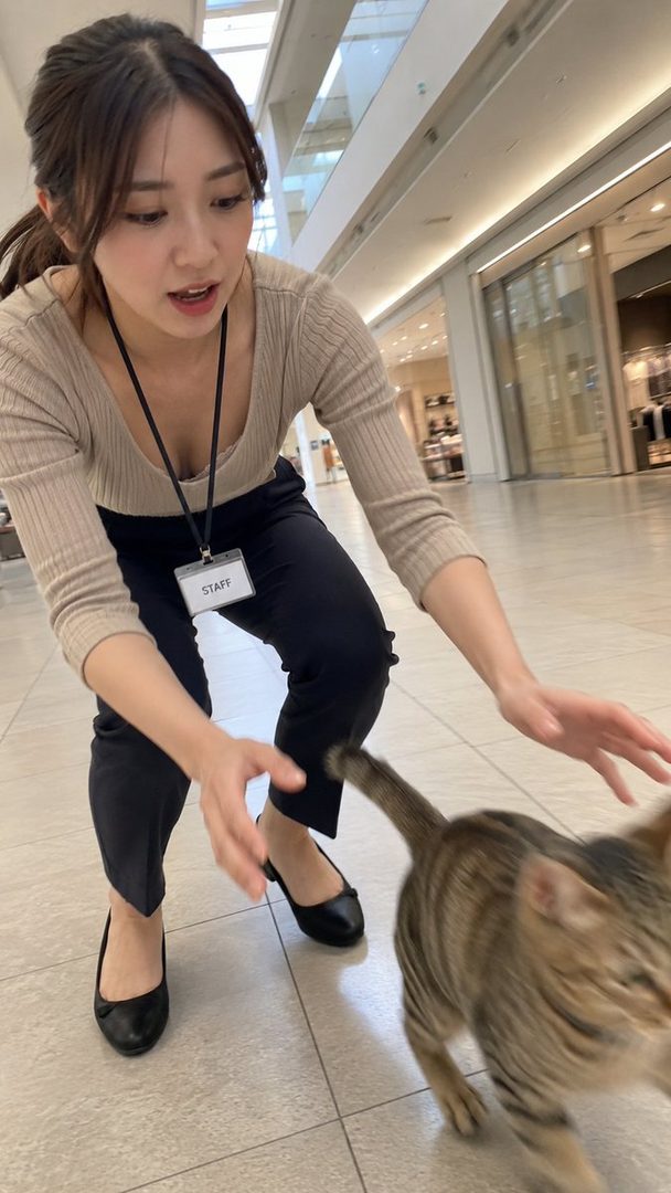 A photorealistic image of a Japanese woman with brown hair, wearing a beige top and black pants, bending down to play with a cat in an indoor shopping mall.