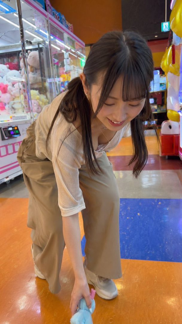 A young Japanese woman with brown hair in pigtails bends down to pick up a blue plush toy in an arcade.