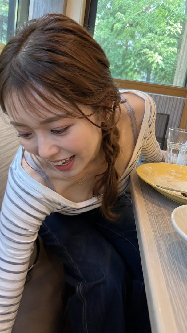 A young woman with brown hair in a braid, wearing a striped top and jeans, smiles while looking down in an indoor setting.